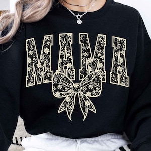 Könnte beinhalten: Schwarzer Pullover mit einem floralen Design, das "MAMA" buchstabiert, mit einer floralen Schleife darunter.