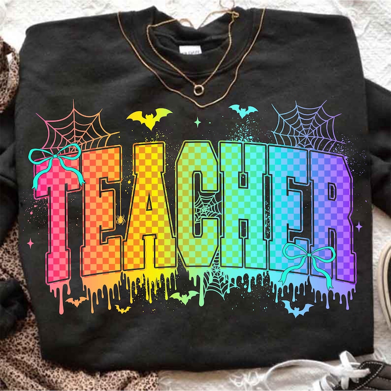 May include: Black sweatshirt with the word "TEACHER" in a rainbow checkered design. The design includes spiderwebs, bats, and bows, with a dripping paint effect at the bottom. A gold necklace is draped over the sweatshirt.