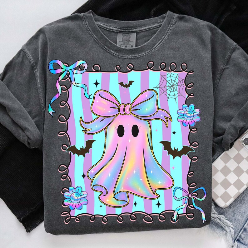 Puede incluir: Una sudadera gris oscuro con un dise&ntilde;o de fantasma pastel. El fantasma tiene un gran lazo y est&aacute; rodeado de murci&eacute;lagos, flores y una telara&ntilde;a. El fondo tiene rayas verticales azules y moradas.
