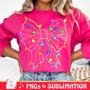 May include: A pink sweatshirt with a white outline of a bow made of Christmas lights. The lights are multi-colored and include red, yellow, green, blue, and purple.