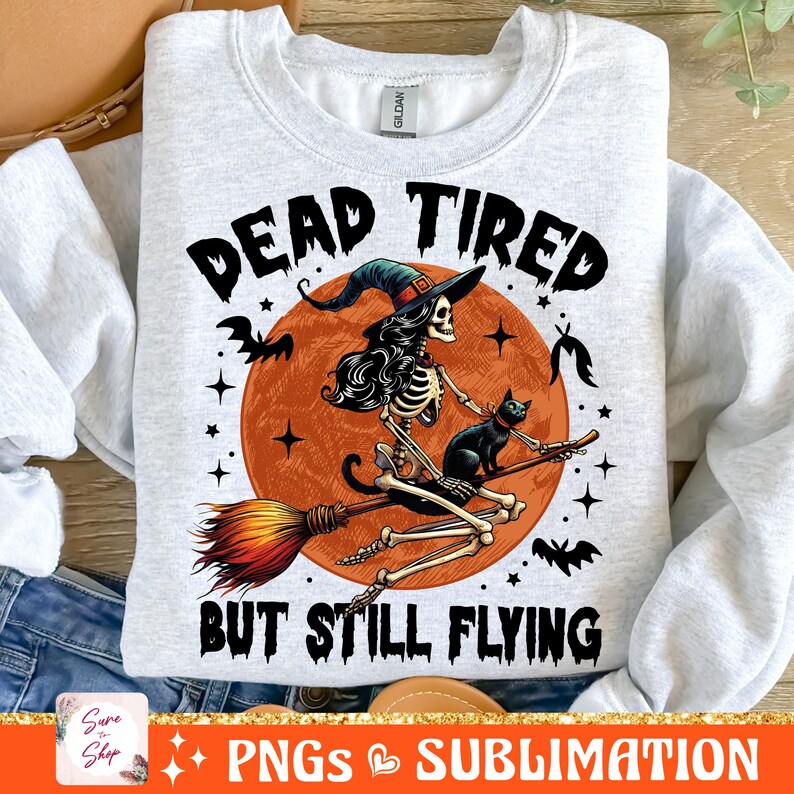May include: A white sweatshirt with a graphic print of a skeleton witch riding a broomstick with a black cat. The text on the sweatshirt reads "Dead Tired But Still Flying".