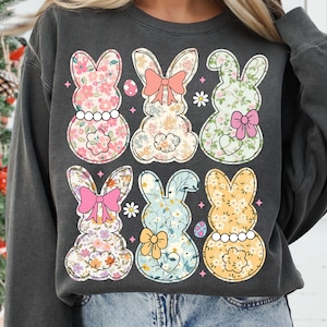 May include: A grey sweatshirt featuring a floral pattern of six Easter bunnies. The bunnies are all different colours and have bows on their ears.