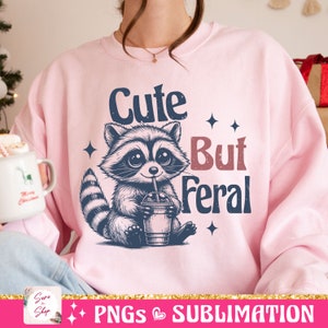 May include: A light pink sweatshirt featuring a raccoon illustration drinking from a cup with a straw. The text "Cute But Feral" is above and to the right of the raccoon. Four small star shapes are also on the sweatshirt.