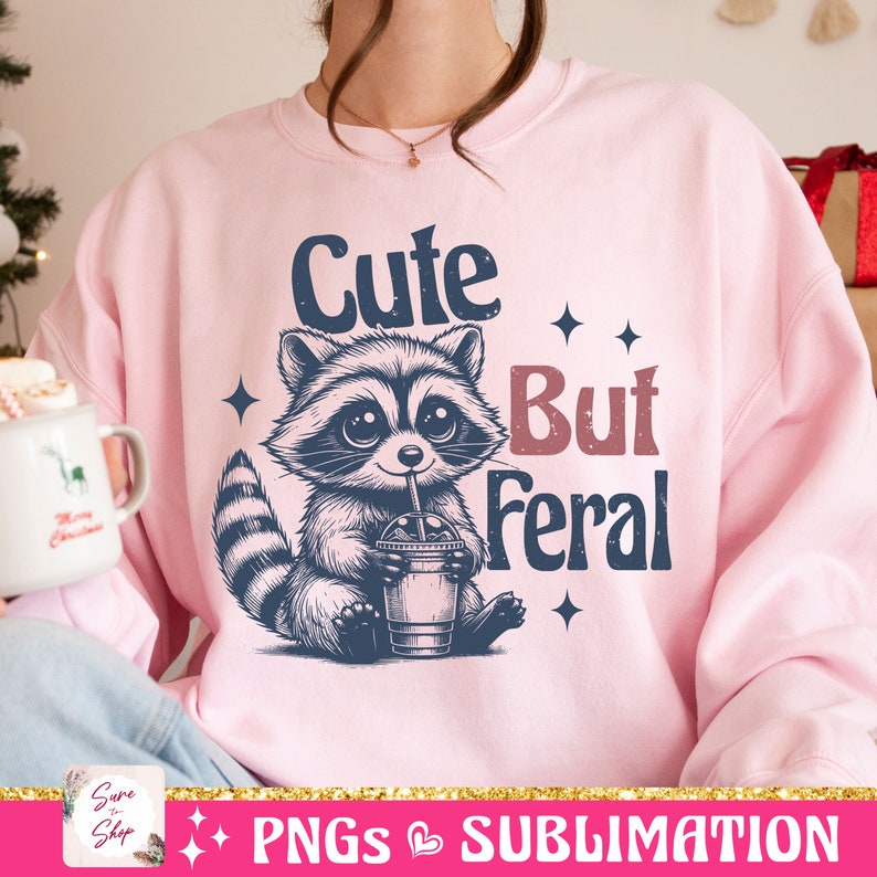 May include: A light pink sweatshirt featuring a raccoon illustration drinking from a cup with a straw. The text "Cute But Feral" is above and to the right of the raccoon. Four small star shapes are also on the sweatshirt.