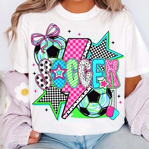 Puede incluir: Camiseta blanca con un dise&ntilde;o gr&aacute;fico de la palabra "SOCCER" en letras de colores con un rayo rosa, estrellas y balones de f&uacute;tbol.