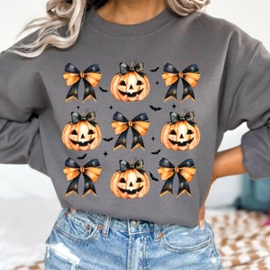 May include: A gray sweatshirt with a repeating pattern of orange jack-o'-lantern pumpkins with black eyes and a toothy grin, each pumpkin is adorned with a black and gold bow.
