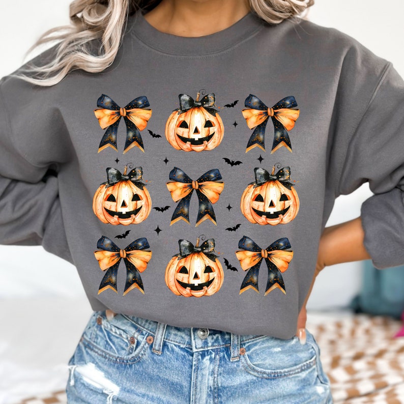 May include: A gray sweatshirt with a repeating pattern of orange jack-o'-lantern pumpkins with black eyes and a toothy grin, each pumpkin is adorned with a black and gold bow.