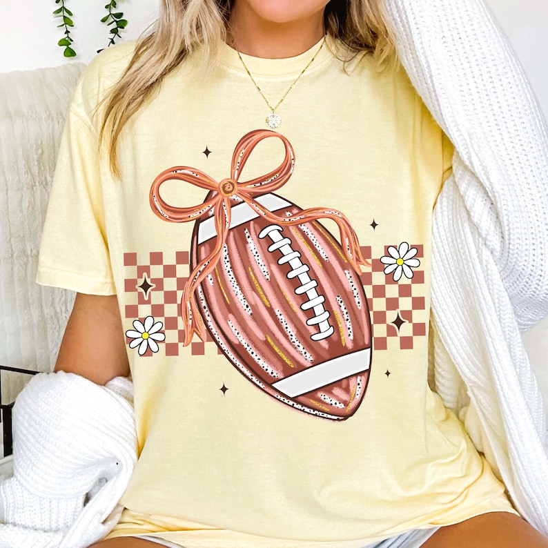 May include: A light yellow t-shirt featuring a football design with a bow. The football is brown with white laces and a white stripe. The design includes daisies, stars, and a checkered pattern in brown and white.