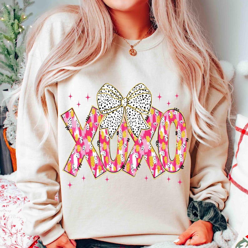K&ouml;nnte beinhalten: Beiges Sweatshirt mit den Buchstaben "XOXO" in einem rosa-gelben Muster und einer wei&szlig;en Schleife mit schwarzen Punkten. Rosa Sternschnuppen umgeben das Design.