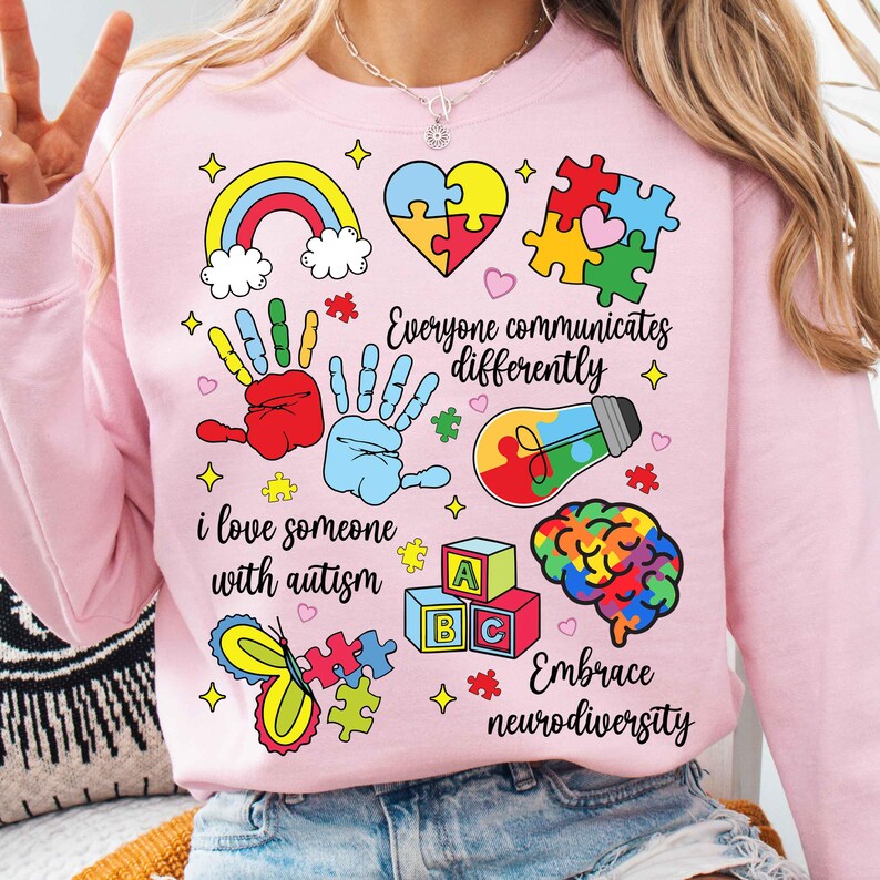 May include: Pink sweatshirt with a colorful design featuring puzzle pieces, handprints, a rainbow, a lightbulb, and the text "Everyone communicates differently", "I love someone with autism", and "Embrace neurodiversity".