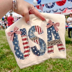 Op de afbeelding: Cr&egrave;mekleurige stof met het woord "USA" in denim, rood-wit gestreepte stof en luipaardprint. Elke letter is versierd met een rood-wit-blauwe gestreepte strik. De afbeelding wordt buiten gehouden.