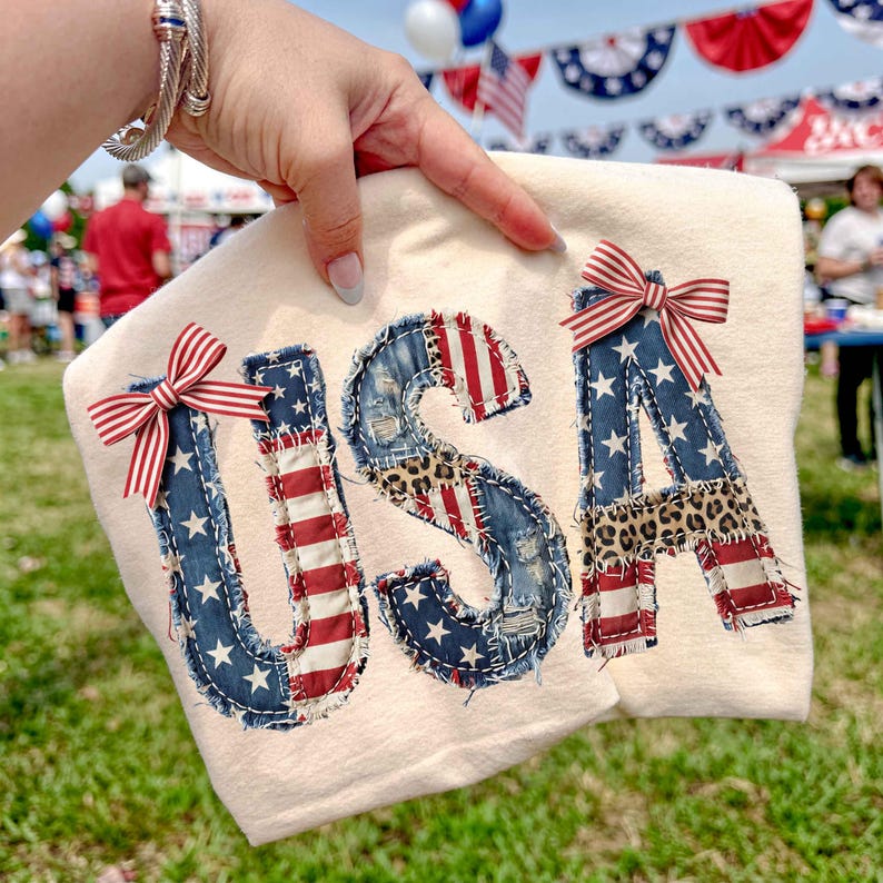 Op de afbeelding: Cr&egrave;mekleurige stof met het woord "USA" in denim, rood-wit gestreepte stof en luipaardprint. Elke letter is versierd met een rood-wit-blauwe gestreepte strik. De afbeelding wordt buiten gehouden.