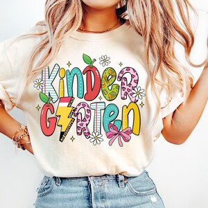 May include: A white t-shirt with a colorful graphic that says "Kindergarten" with a variety of patterns and designs, including hearts, stars, and a lightning bolt.