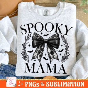 May include: A white sweatshirt with the text "Spooky Mama" in black lettering. A black lace bow with a black ribbon is centered on the sweatshirt. The bow is surrounded by a wreath of leaves and bats.