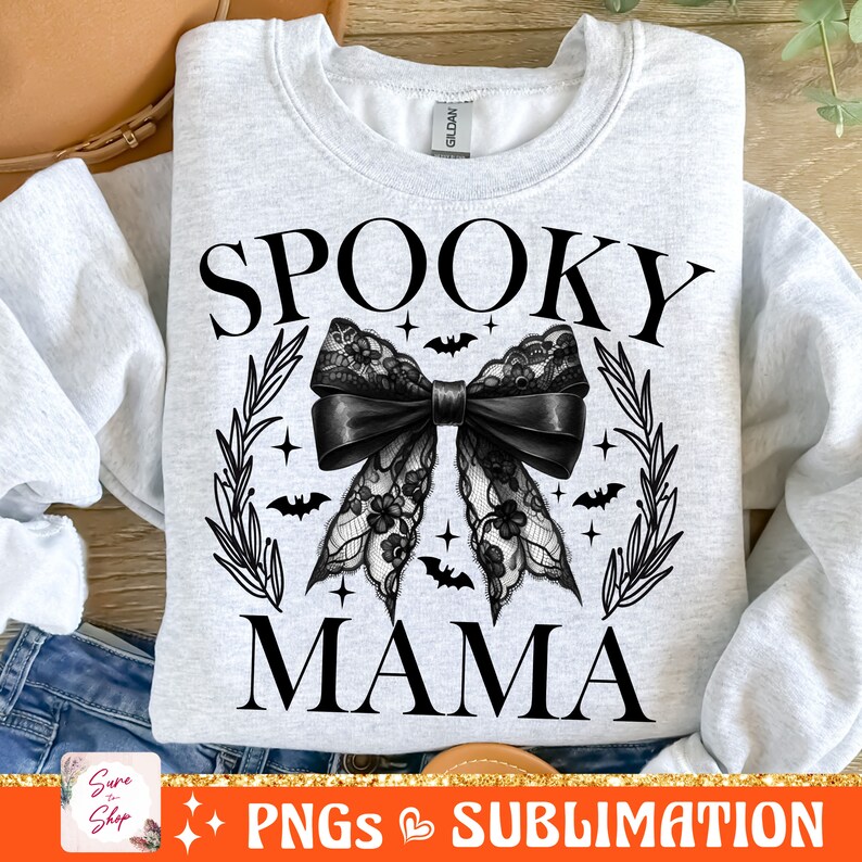May include: A white sweatshirt with the text "Spooky Mama" in black lettering. A black lace bow with a black ribbon is centered on the sweatshirt. The bow is surrounded by a wreath of leaves and bats.