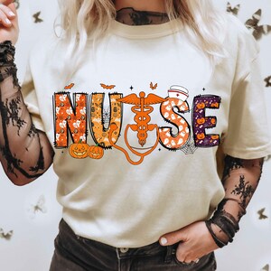 May include: Cream-colored t-shirt with the word "NURSE" in a Halloween-themed design. The letters are filled with pumpkins, ghosts, and spiderwebs. An orange caduceus and stethoscope are featured in the design.