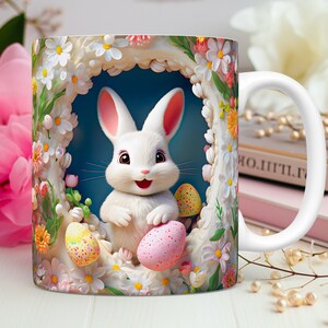 May include: White ceramic mug featuring a colourful Easter design. A white bunny rabbit holds two pink and yellow speckled Easter eggs. The bunny is surrounded by white and pink flowers.
