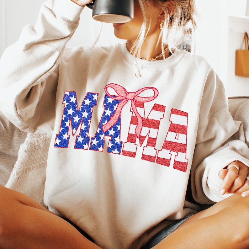 May include: A white sweatshirt with a red, white, and blue "MAMA" graphic. The letters are decorated with stars and stripes, and a pink bow is centered above the word.