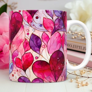 May include: White ceramic coffee mug featuring a vibrant design of stylized pink and purple leaves with gold outlines. The mug has a white handle and is set against a soft background with a pink flower.