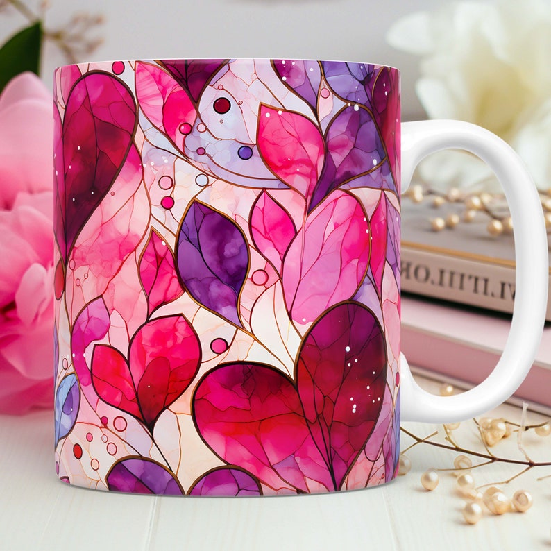 May include: White ceramic coffee mug featuring a vibrant design of stylized pink and purple leaves with gold outlines. The mug has a white handle and is set against a soft background with a pink flower.