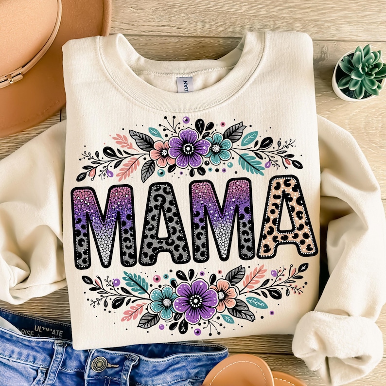 May include: A cream-coloured sweatshirt with the word "MAMA" in black letters with a leopard print and purple, pink, and blue glitter. The word is surrounded by a floral design with pink, purple, and blue flowers.