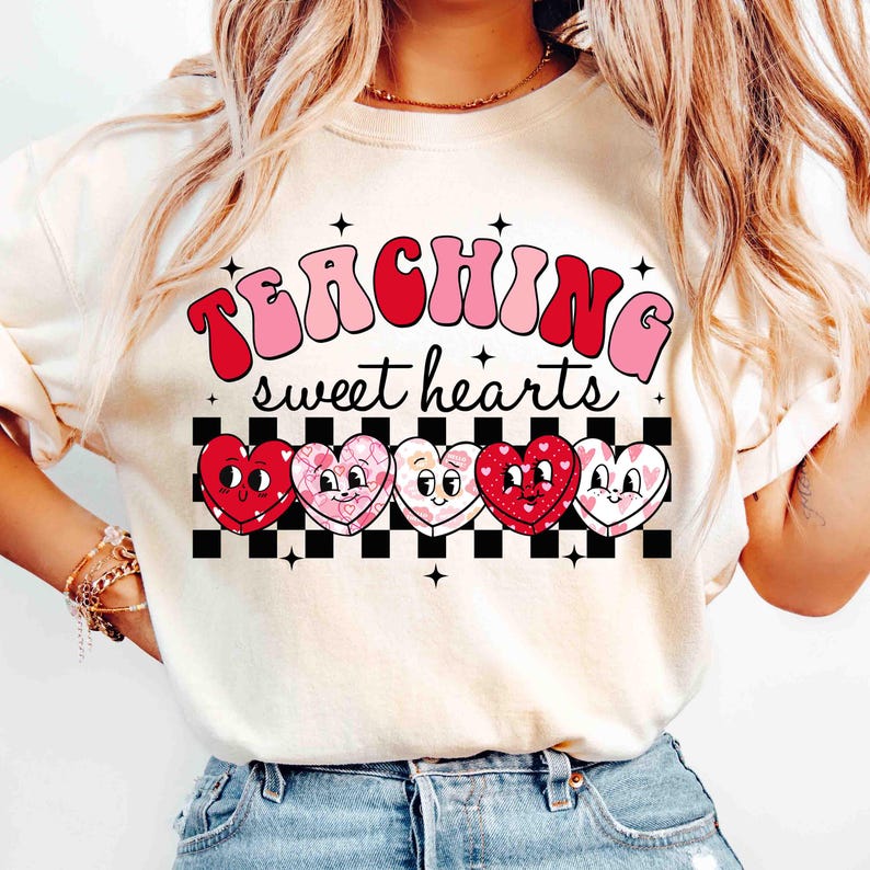 Pu&ograve; includere: Una maglietta bianca con il testo "TEACHING sweet hearts" in lettere rosa e rosse. Il testo &egrave; circondato da un motivo a scacchi e cinque cuori di cartoni animati con accenti rosa e rossi.