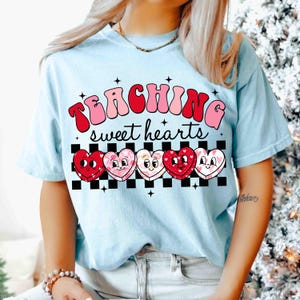 Pu&ograve; includere: Maglietta blu chiaro con il testo "TEACHING sweet hearts" in lettere rosa e rosse. Cinque cuori di cartone animato con facce sono allineati sotto il testo su uno sfondo a scacchi bianco e nero.