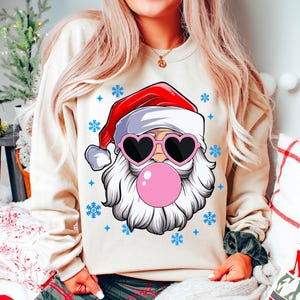 May include: A cream-colored sweatshirt with a graphic of Santa Claus wearing heart-shaped sunglasses and blowing a pink bubble. The graphic is surrounded by blue snowflakes.