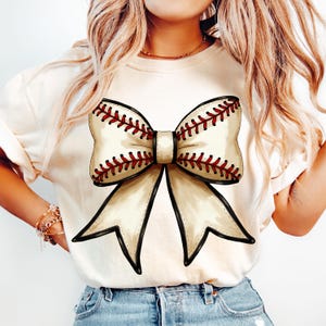 Puede incluir: Una camiseta blanca con un gran lazo de b&eacute;isbol en crema y rojo. El lazo est&aacute; atado en un nudo y los extremos caen hacia abajo.