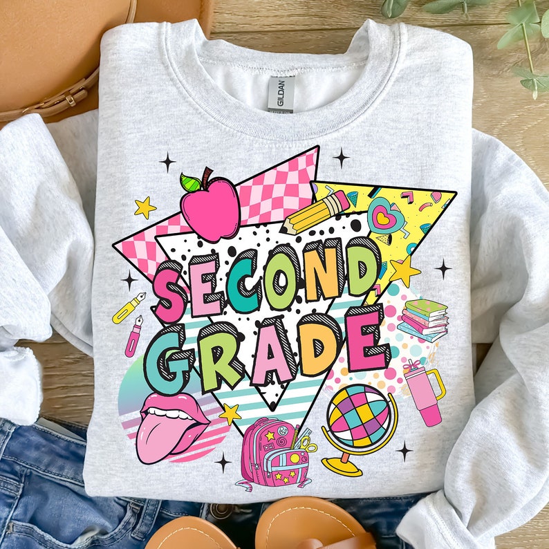 K&ouml;nnte beinhalten: Ein graues Sweatshirt mit einem bunten Grafikdesign, das die Worte "Second Grade" im Retro-Stil zeigt. Das Design beinhaltet einen roten Apfel, einen Bleistift, einen Globus, einen Rucksack und andere schulthemenbezogene Elemente.