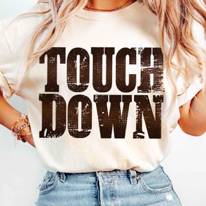 May include: Cream-colored sweatshirt with the words "TOUCH DOWN" printed in a distressed, dark brown font. The text has a textured, vintage appearance. The shirt is a casual, everyday wear item.