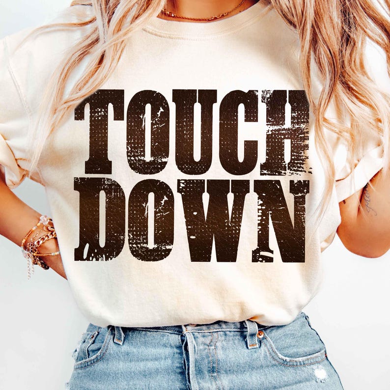May include: Cream-colored sweatshirt with the words "TOUCH DOWN" printed in a distressed, dark brown font. The text has a textured, vintage appearance. The shirt is a casual, everyday wear item.