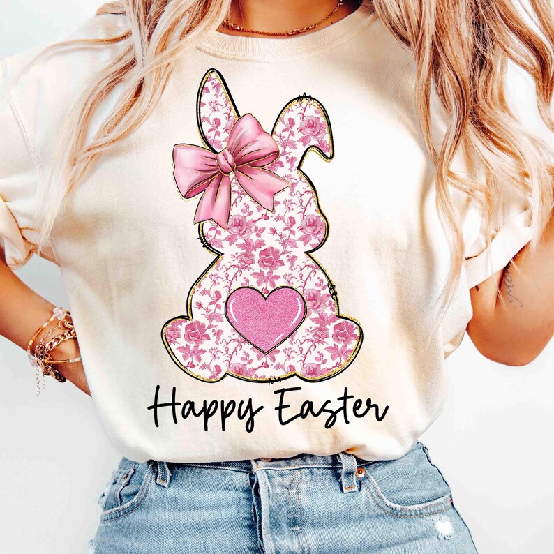 Op de afbeelding: Een wit T-shirt met een roze bloemachtig konijnenontwerp en een roze strik. Het konijn heeft een roze hart in het midden van zijn borst. De tekst "Happy Easter" is onder het konijn gedrukt.