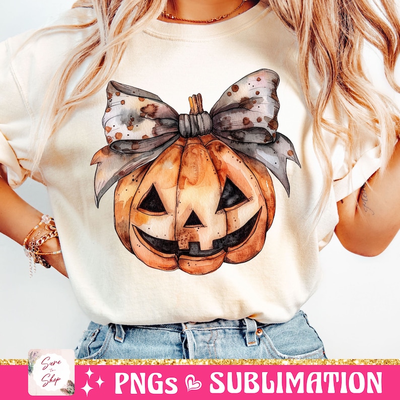 May include: A white t-shirt with a watercolor illustration of a pumpkin with a brown and gray bow. The pumpkin has a carved jack-o'-lantern face.