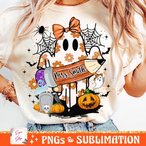 Puede incluir: Una camiseta blanca con un fantasma sosteniendo un l&aacute;piz que dice "Mrs. Smith". El fantasma est&aacute; decorado con flores naranjas y est&aacute; rodeado de elementos de tem&aacute;tica de Halloween como una telara&ntilde;a, murci&eacute;lagos y calabazas.