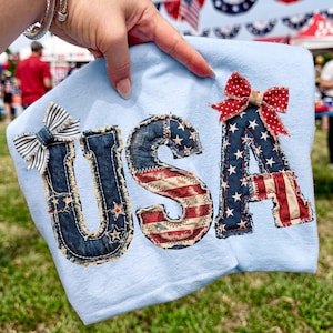 May include: Light blue shirt with the letters "USA" appliqued in patriotic colors and patterns. The letters are embellished with stars and stripes, and the "U" has a striped bow. The "A" has a red polka dot bow.