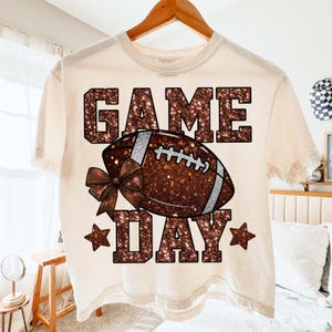 Puede incluir: Camiseta color crema con un dise&ntilde;o tem&aacute;tico de f&uacute;tbol americano. El dise&ntilde;o presenta las palabras "GAME DAY" en una fuente marr&oacute;n brillante, un bal&oacute;n de f&uacute;tbol marr&oacute;n con detalles blancos, un lazo y estrellas. La camiseta est&aacute; colgada en una percha de madera.