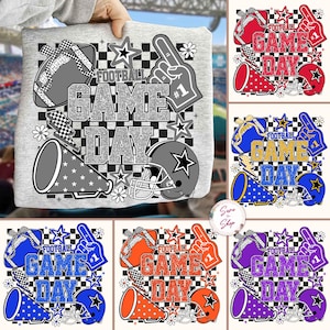 May include: A grey t-shirt featuring a football-themed design. The design includes a football, a helmet, a foam finger, and the words "FOOTBALL GAME DAY" in a glittery font. Additional designs include stars, flowers, and a checkered pattern.