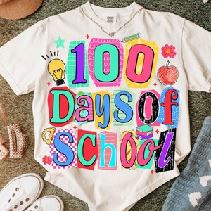 Puede incluir: Camiseta color crema con un gr&aacute;fico colorido que dice "100 Days of School". El dise&ntilde;o incluye una bombilla, una manzana, libros y otros elementos escolares en colores brillantes. La camiseta est&aacute; hecha de un material suave.