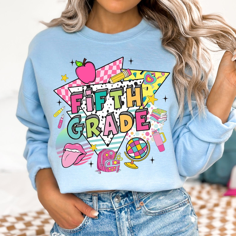 K&ouml;nnte beinhalten: Ein hellblaues Sweatshirt mit einem grafischen Design, das die Worte "FIFTH GRADE" in einem farbenfrohen Retro-Stil zeigt. Das Design beinhaltet einen roten Apfel, einen Bleistift, einen Globus, einen Rucksack und andere schulthemenbezogene Elemente.