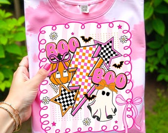 Puede incluir: Camiseta rosa teñida con un diseño de Halloween. El diseño incluye la palabra "BOO" en letras rosas, una calabaza, un fantasma, estrellas y murciélagos. La camiseta tiene cuello redondo y mangas cortas.