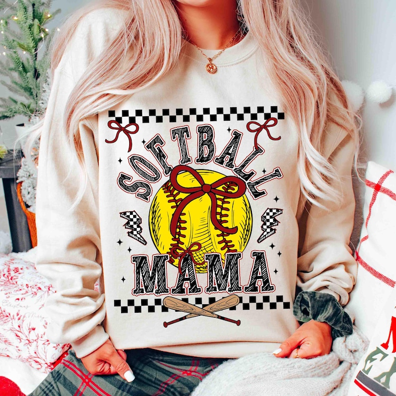 May include: Cream-colored sweatshirt with the words "SOFTBALL MAMA" in black and red lettering. A yellow softball with a red bow is in the center. Two crossed baseball bats are at the bottom. Checkered patterns and lightning bolt graphics complete the design.