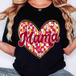 K&ouml;nnte beinhalten: Schwarzes T-Shirt mit einem herzf&ouml;rmigen Design. Das Herz hat ein rot-wei&szlig;es Karomuster, goldene Akzente und kleine rote Herzen. Das Wort "Mama" ist in roter Schrift &uuml;ber dem Herzen geschrieben.