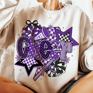 May include: Cream-colored sweatshirt with a large, colorful "Cheer" graphic. The design features purple, black, and white elements, including stars, pom-poms, and a megaphone. The word "Cheer" is in a bold, playful font, perfect for cheerleading.