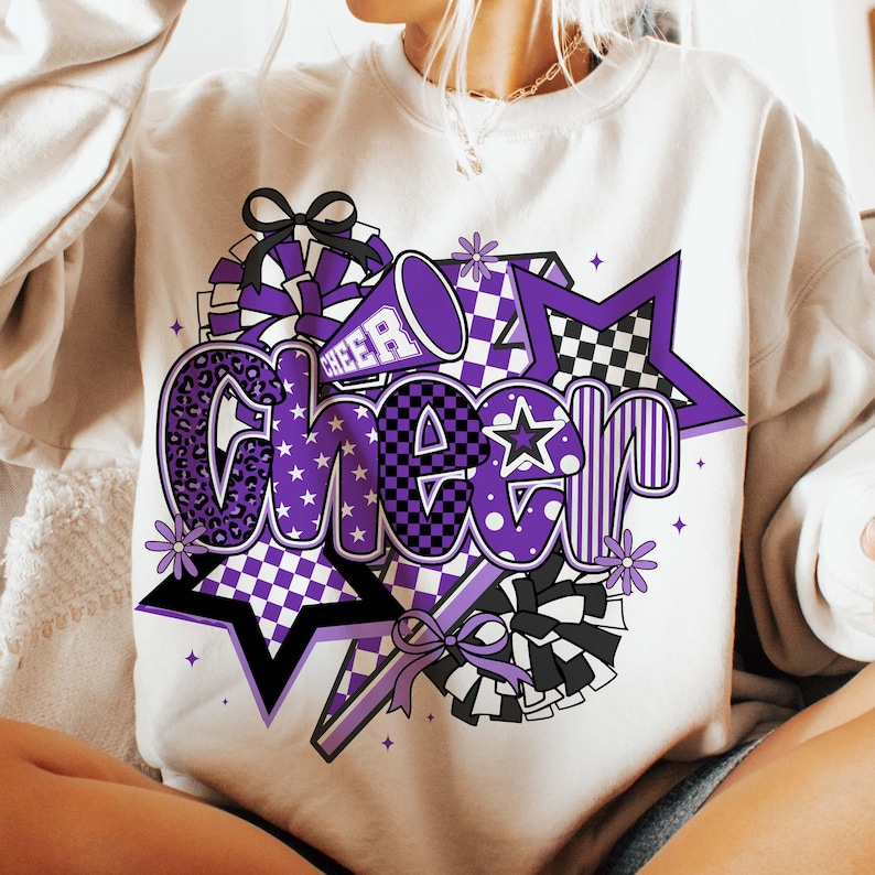 May include: Cream-colored sweatshirt with a large, colorful "Cheer" graphic. The design features purple, black, and white elements, including stars, pom-poms, and a megaphone. The word "Cheer" is in a bold, playful font, perfect for cheerleading.