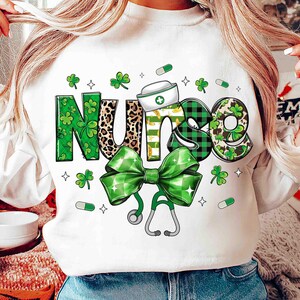 Pu&ograve; includere: Felpa bianca con la scritta "Nurse" in un design decorativo. Le lettere sono decorate con trifogli, stampa leopardata e motivi a quadri verdi e neri. Sono inclusi anche un fiocco verde e uno stetoscopio.