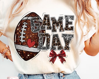 May include: Cream-colored t-shirt featuring a football graphic with the words "GAME DAY" in a combination of leopard print and glitter. A red bow is below the text, and a heart is on the football.