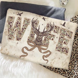 May include: A rectangular pillow with the word "WIFE" in camouflage print. A deer head graphic is in the center, with a brown ribbon below. The pillow has a vintage, distressed look, with small bird and star graphics.