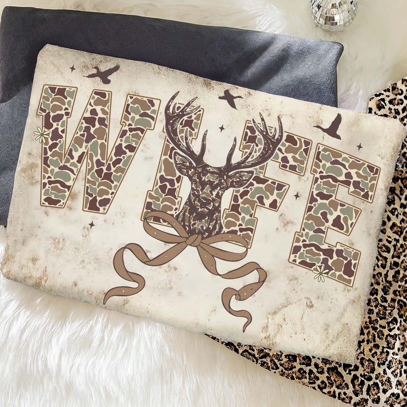 May include: A rectangular pillow with the word "WIFE" in camouflage print. A deer head graphic is in the center, with a brown ribbon below. The pillow has a vintage, distressed look, with small bird and star graphics.
