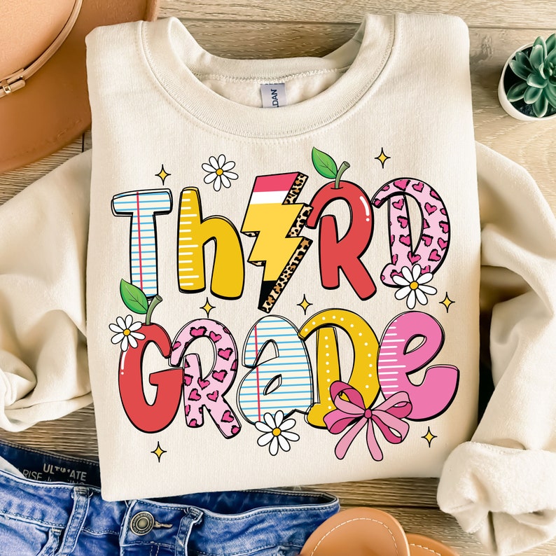 May include: A beige sweatshirt with the words "Third Grade" in colorful, playful letters. The letters are decorated with hearts, stars, and other school-themed designs.